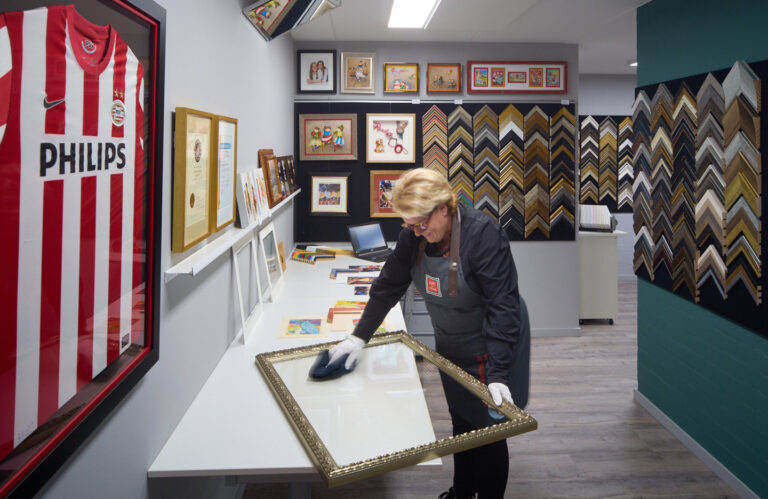 De showroom van Heijnen Decor met een ingelijst PSV-shirt, een wand vol ingelijste werken en honderden lijstprofielen, terwijl Natascha het glas van een vergulde lijst schoonmaakt